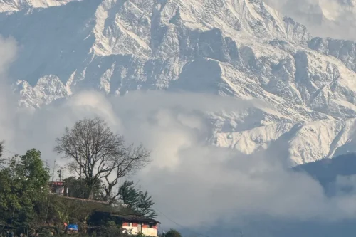 Short Hikes Near Kathmandu featuring a scenic hillside village beneath snow-capped Himalayan peaks, with clouds drifting across the mountains and lush greenery in the foreground.