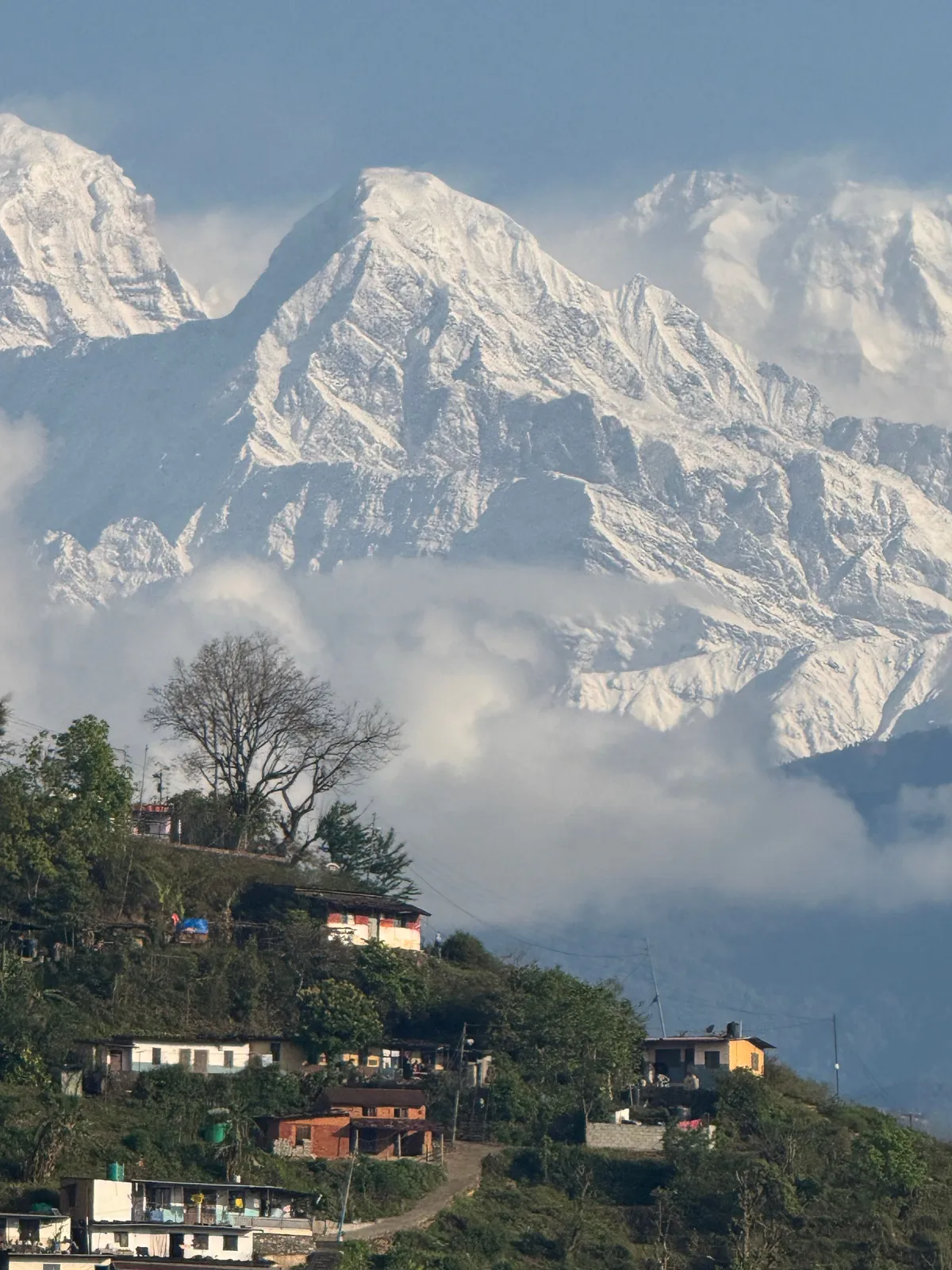 Short Hikes Near Kathmandu featuring a scenic hillside village beneath snow-capped Himalayan peaks, with clouds drifting across the mountains and lush greenery in the foreground.