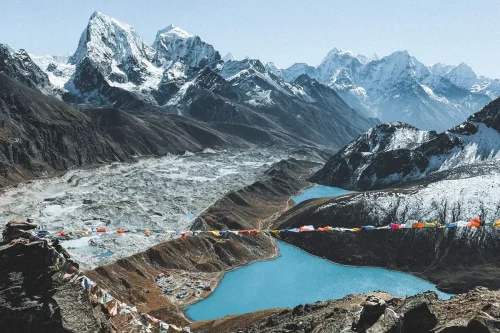 Best Treks in Nepal featuring a stunning Himalayan mountain landscape, turquoise glacial lake, glacier, rocky terrain, and colorful prayer flags.