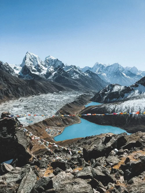 Best Treks in Nepal featuring a stunning Himalayan mountain landscape, turquoise glacial lake, glacier, rocky terrain, and colorful prayer flags.